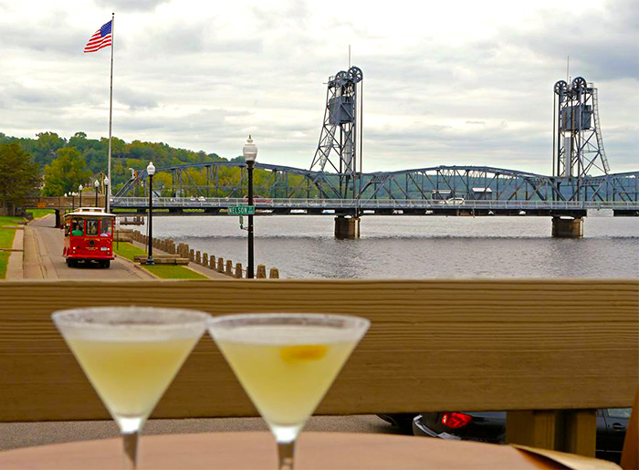 A bridge spans a body of water and ends beside a flagpole with the American flag and a trolley car running nearby, with a pair of mixed drinks in martini glasses in the foreground in Stillwater, MN.